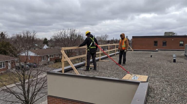 Couvreur en harnais relié à un ancrage lors d'un travail en hauteur sur un toit au Québec, avec garde-corps et collègue en surveillance