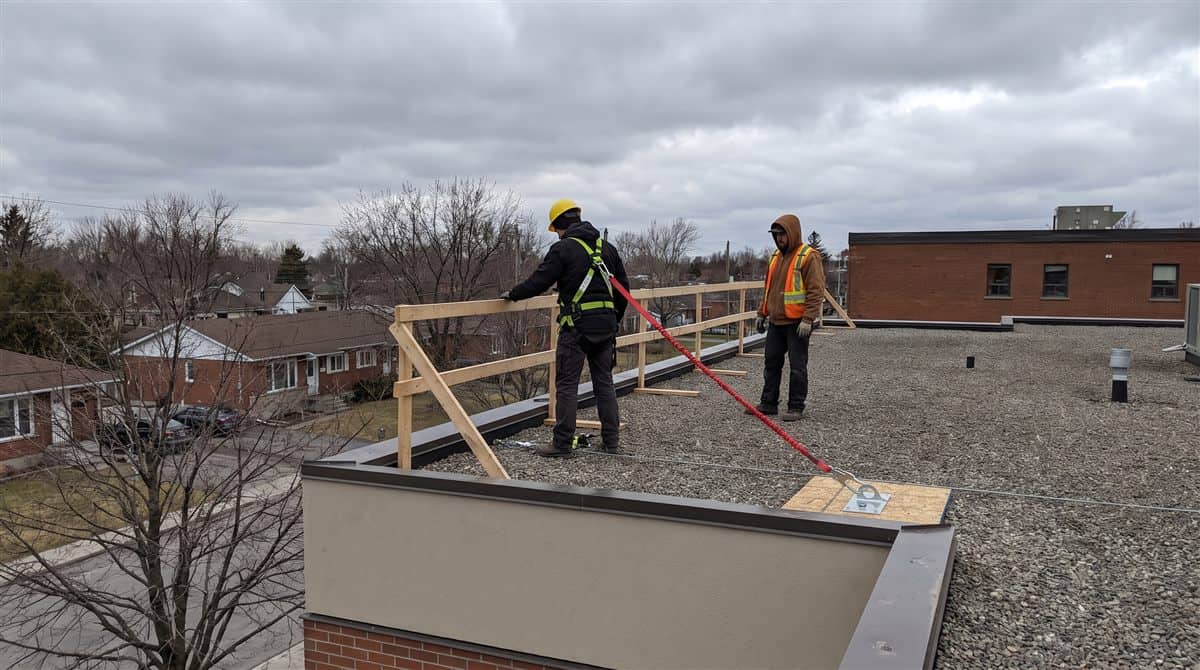 Couvreur en harnais relié à un ancrage lors d'un travail en hauteur sur un toit au Québec, avec garde-corps et collègue en surveillance