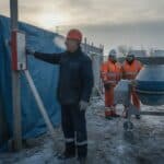 Chef d'entreprise de maçonnerie qui montre un thermomètre sous zéro sur un chantier givré, deux ouvriers en gilet orange qui attendent près d'une bétonnière à l'arrêt