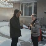 Entrepreneur en manteau d'hiver remet un papier à une femme âgée souriante devant un bungalow enneigé, souffleuse et pelle dans l'entrée déneigée